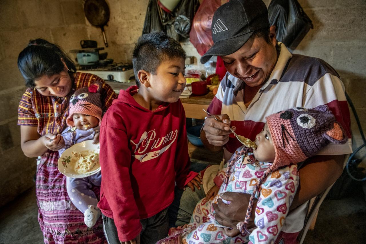 A mother and father feed their infant twins, while their young son looks on and tries to help.