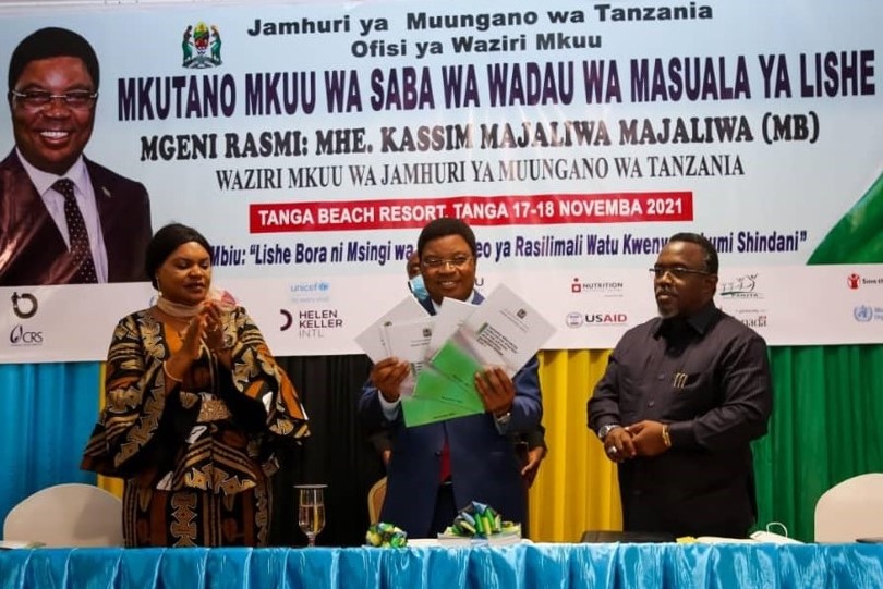 A Tanzanian government official stands and holds up copies of the NMNAP II document, while two other onlookers stand by his right and left side.