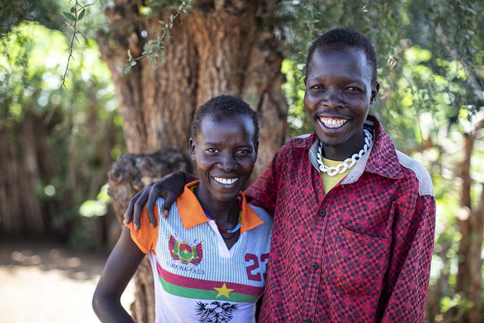 A smiling Ugandan couple stands together outside near a tree lined area.