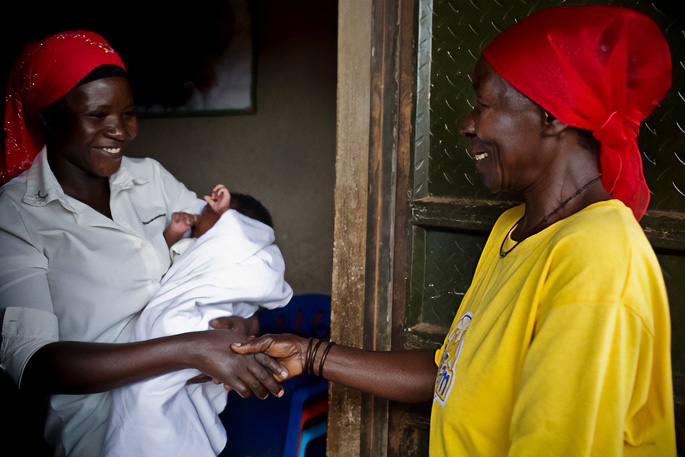 A woman in a bright yellow t-shirt is at the front door of a woman in a white button up shirt, holding her infant child swaddled in a white blanket. The women are shaking hands.