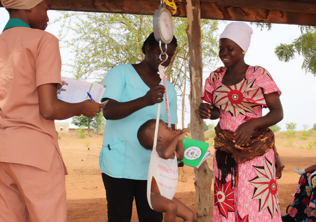 Two Ghanaian Health workers weighing a small child outside as the mother looks on.