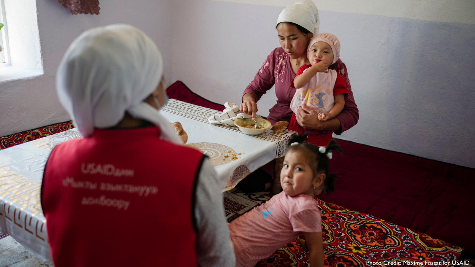Photo of a health worker, talking to a mother holding her small infant, while her toddler sits near her.
