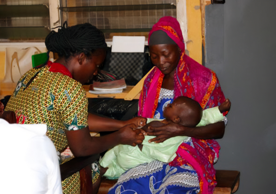 Woman sitting in her house, holding her young infant, as a health worker does a physical check of the child.