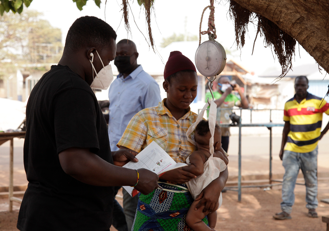 Health worker documenting small infant's weight outside with a hanging scale, while the mother holding the child.