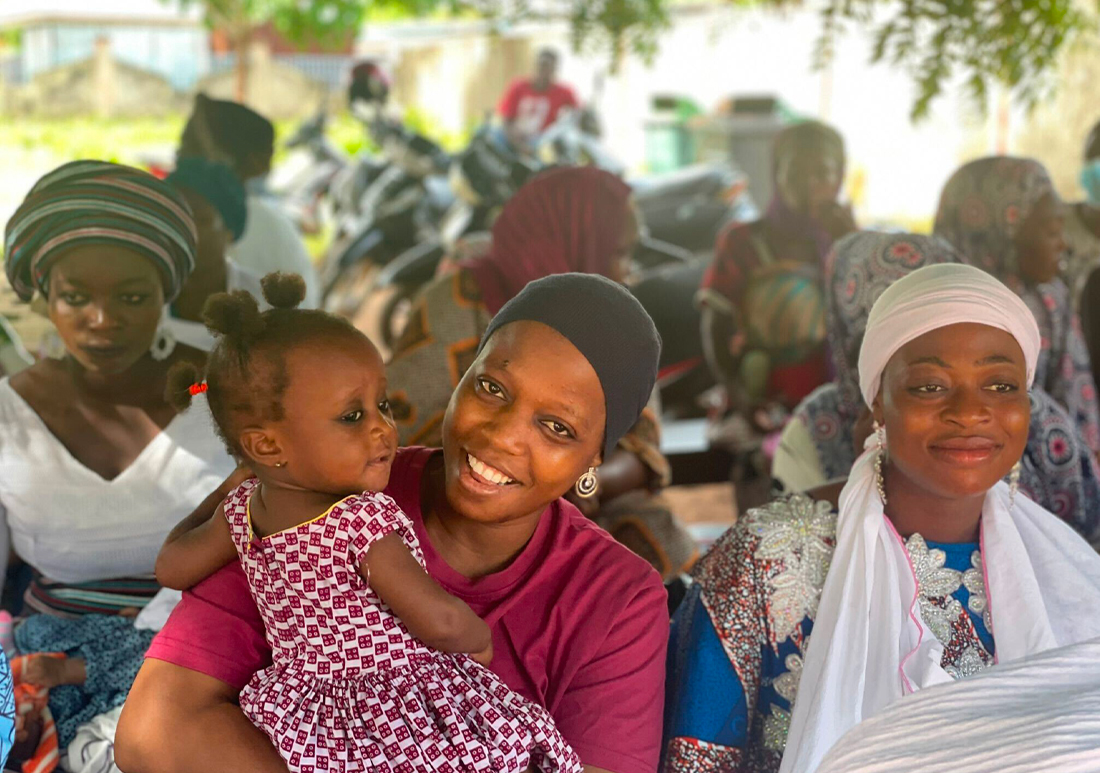 Group of mothers holding their children while sitting under a tree in Ghana. The two mothers in the front of the group are smiling :).