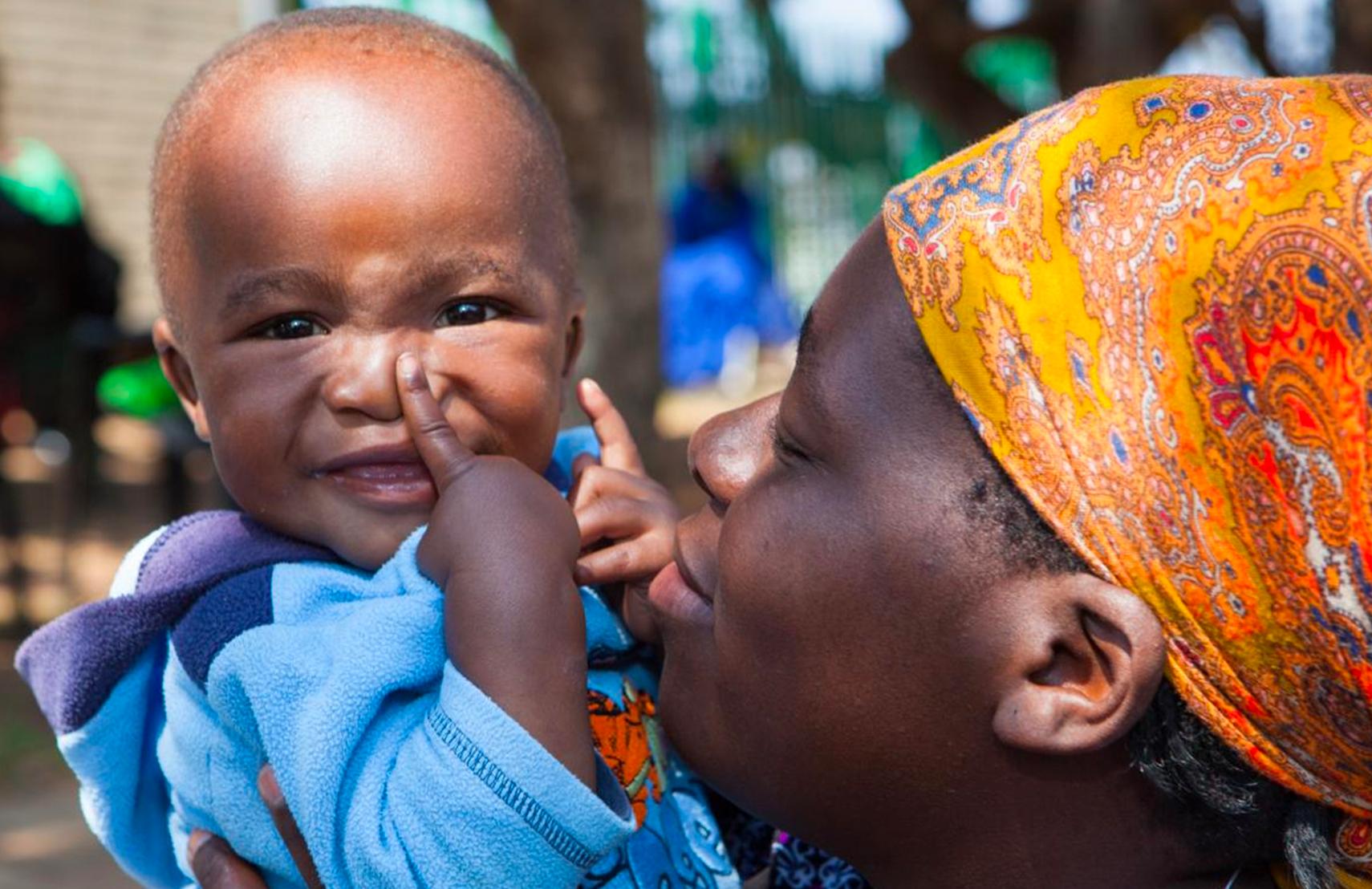 Mother holding her child at a market, and the child is smiling and pointing at their nose.