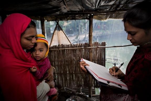 A young mother wearing a red sari is standing up and holding her young infant in her arms, while looking on at a woman that is working as an enumerator and who is writing down data in a notebook 
