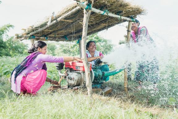 Three female farmers from Nepal in bright saris are outdoors in a field, working to manage a machine used for irrigation.