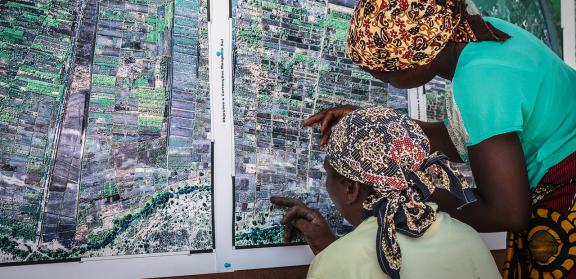Two women are facing a wall-mounted map and are examining points on it. Both women's faces are not visible, but each is wearing a colorful headwrap.
