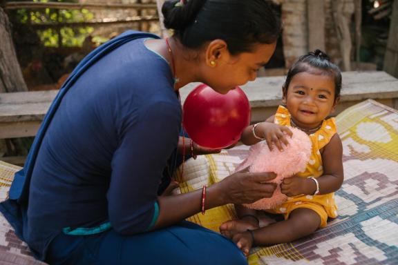 A woman in a blue sari is sitting on the ground with her infant daughter, holding a red ball in her left hand and a plush pink toy in her right, that the girl child is also holding onto with both her hands.