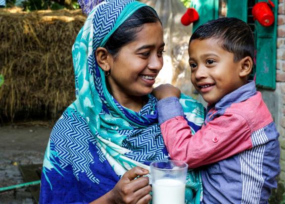 A woman is holding her young son in her left arm and a cup of milk in her right had. She is wearing a blue patterned sari and the boy is wearing a read and blue colored long sleeve shirt.