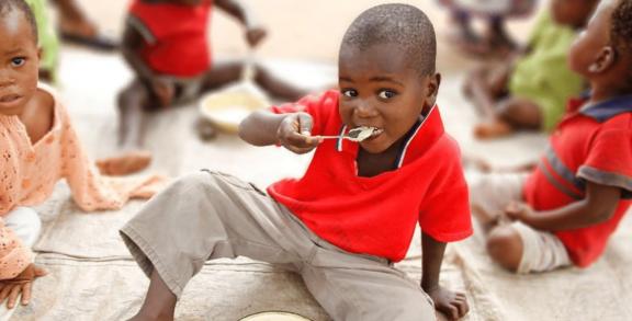 A young boy in a red shirt and brown pants is sitting on the ground eating from a spoon he's holding up to his mouth. Other children are also sitting around him eating.
