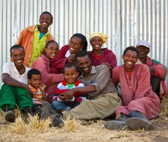 A group of smiling people sit gathered together on the ground outside a building used for agricultural purposes. 