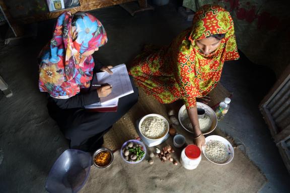 Two women sit on a floor mat measuring various food items; one woman is using a pen and notebook.