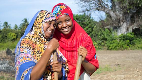 Two young Tanzania women standing together in a field, holding the end of a long farming tool.