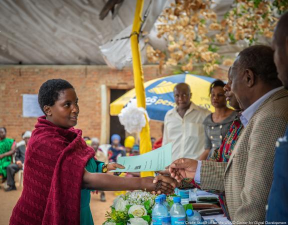 Photo of a woman shacking an older man's hand, while she hands out a document in a public market.