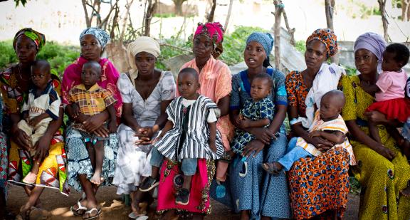 Photo of Rahama, 30 (far right), holds her 9-month-old daughter, Maradiatu, at an Early Childhood Development session that is being run by Mubarak Mohamadu, a Community Health Officer trained in Early Childhood Development. There are also other mothers with their young children as well in this photo.