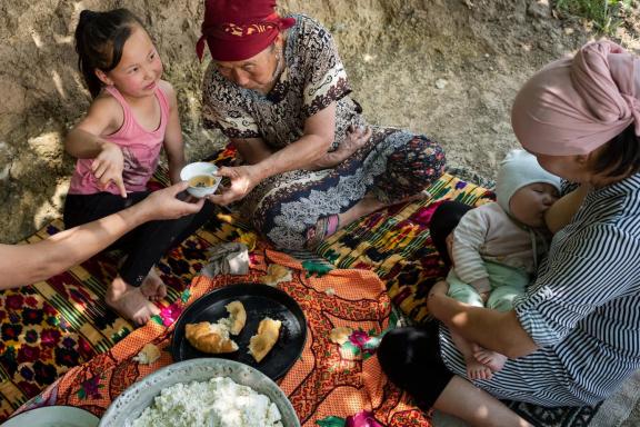 A elderly woman, mother, and children are all gathered sharing a meal on a blanket on the ground.
