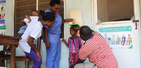 Doctor measuring a young annoyed child's hight, while a nurse and their mother (who is holding a small infant), look on with smiles.