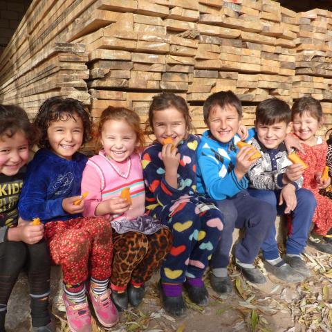 A group of children sit near piles of stacked wooden boards. They are all holding and eating mangos