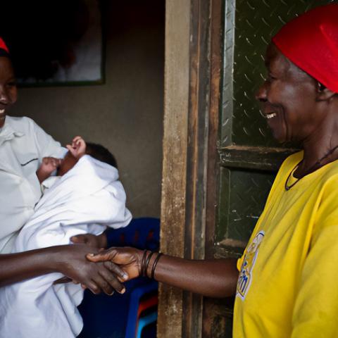 A woman in a bright yellow t-shirt is at the front door of a woman in a white button up shirt, holding her infant child swaddled in a white blanket. The women are shaking hands.