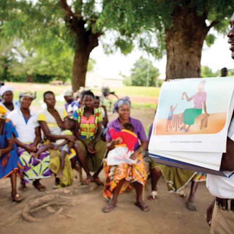 Trainer outside under a tree, showing a group of women an illustration of child play.