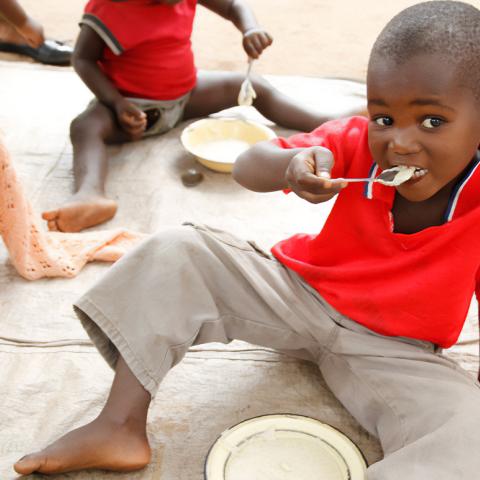 Photo of a small child eating some porridge while sitting down outside, with other children eating around them. 
