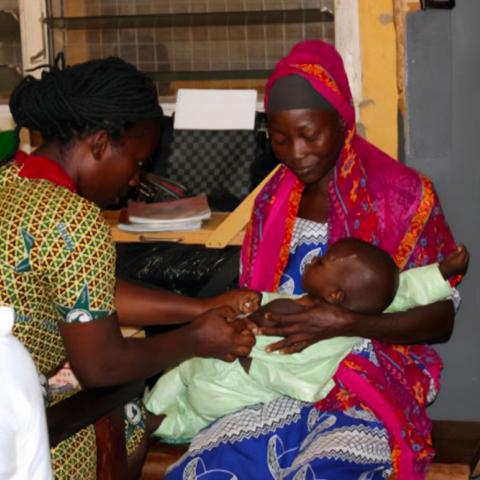 Woman sitting in her house, holding her young infant, as a health worker does a physical check of the child.