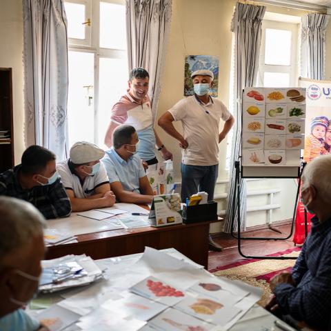 Group of people in a meeting room, wearing masks and reviewing photos of various healthy food on a flip-chart.