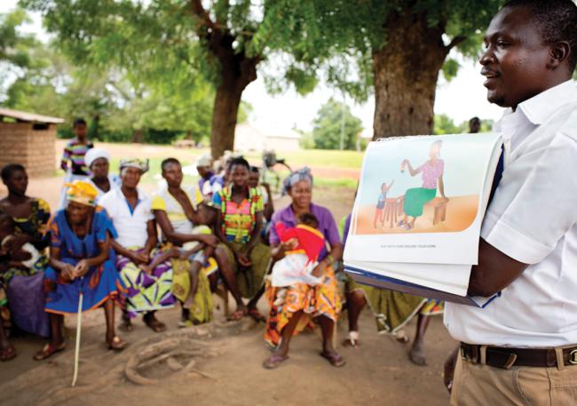 Trainer outside under a tree, showing a group of women an illustration of child play.