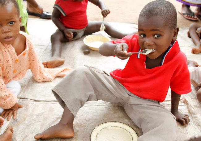 Photo of a small child eating some porridge while sitting down outside, with other children eating around them. 