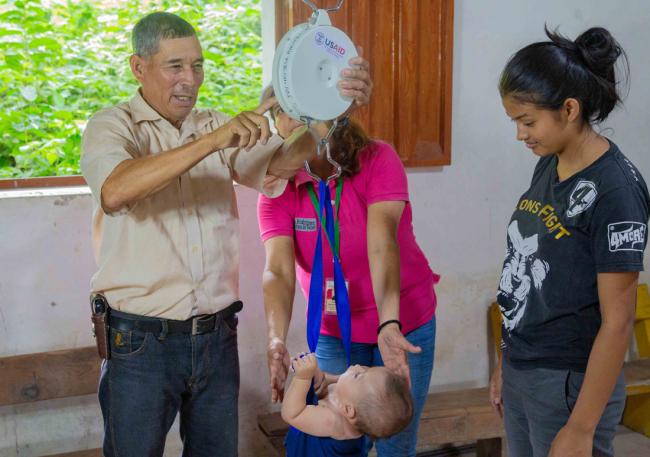 Small infant being weighed by two health professionals, as the mother watches on.