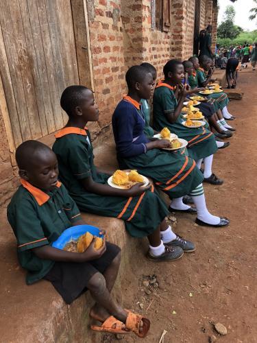 Ugandan students sitting outside in line, eating orange flesh sweet potato. Credit: Feed the Future.